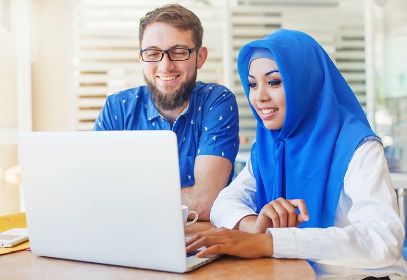 asian muslim woman and caucasian man looking at screen of laptop