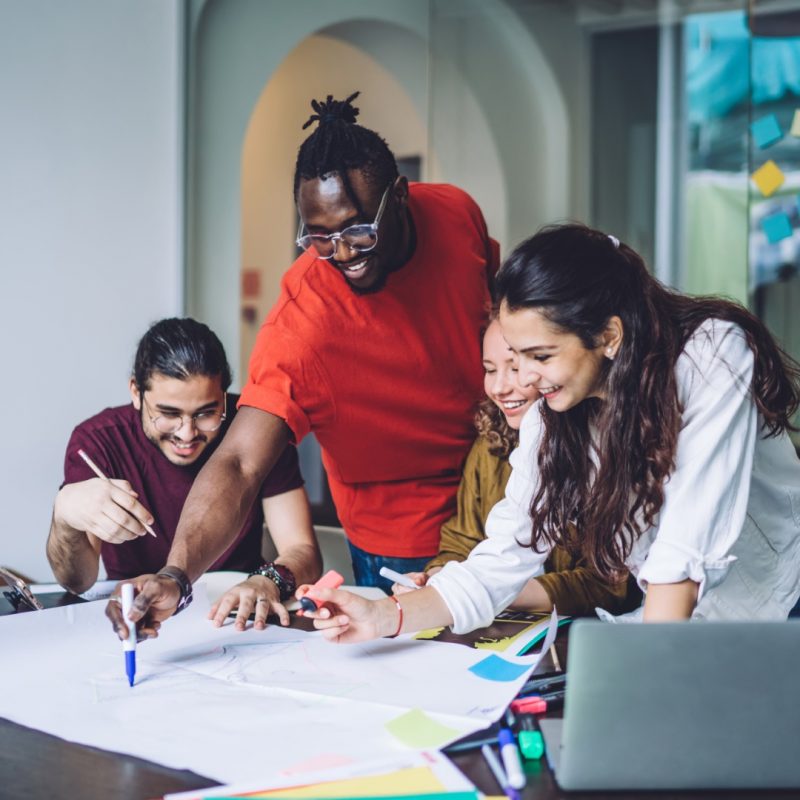 Smiling African American man drawing on paper poster while coworking with diverse group mates and creating new project in team